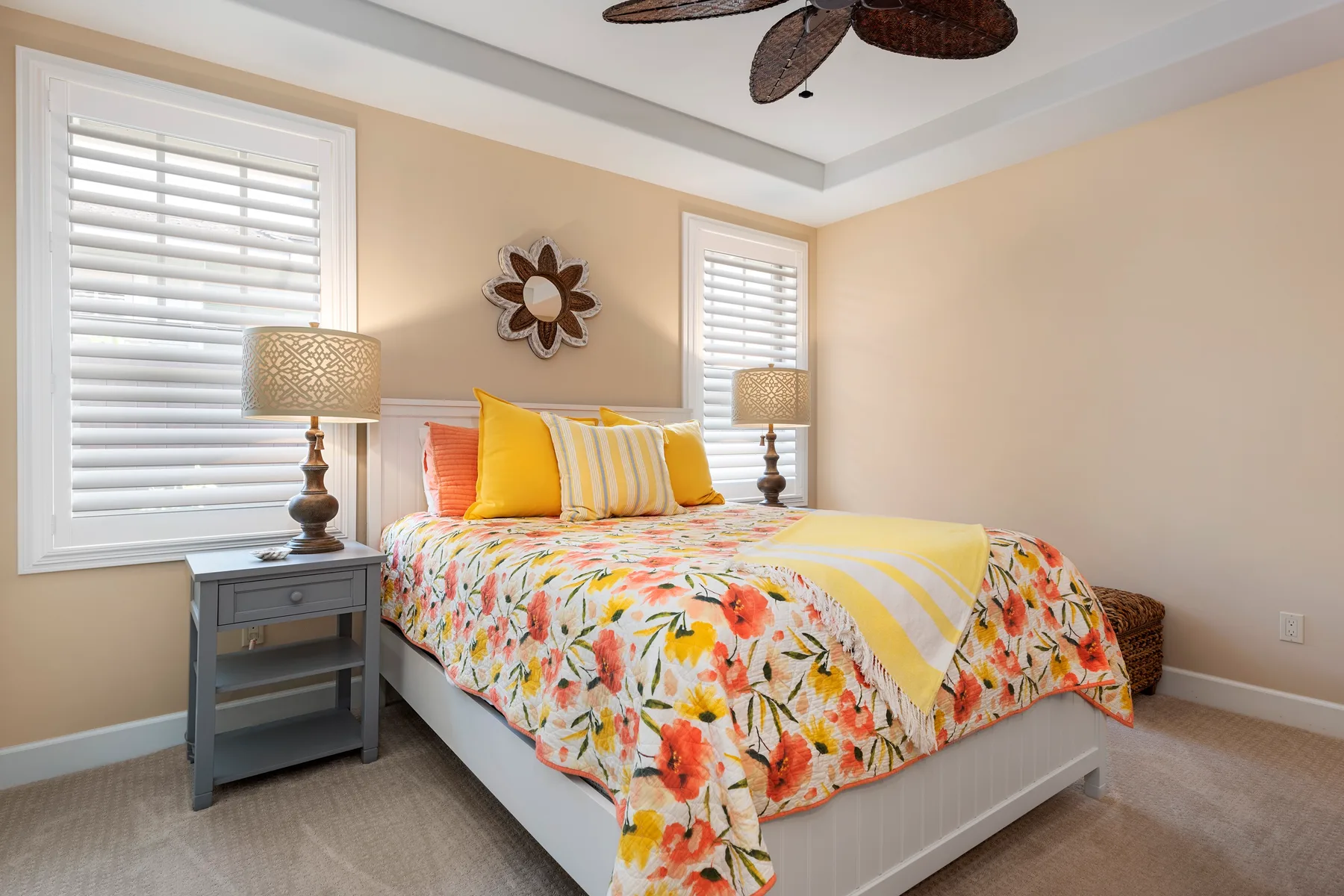 Un dormitorio con una colcha floral, cojines a rayas y ventanas con persianas.