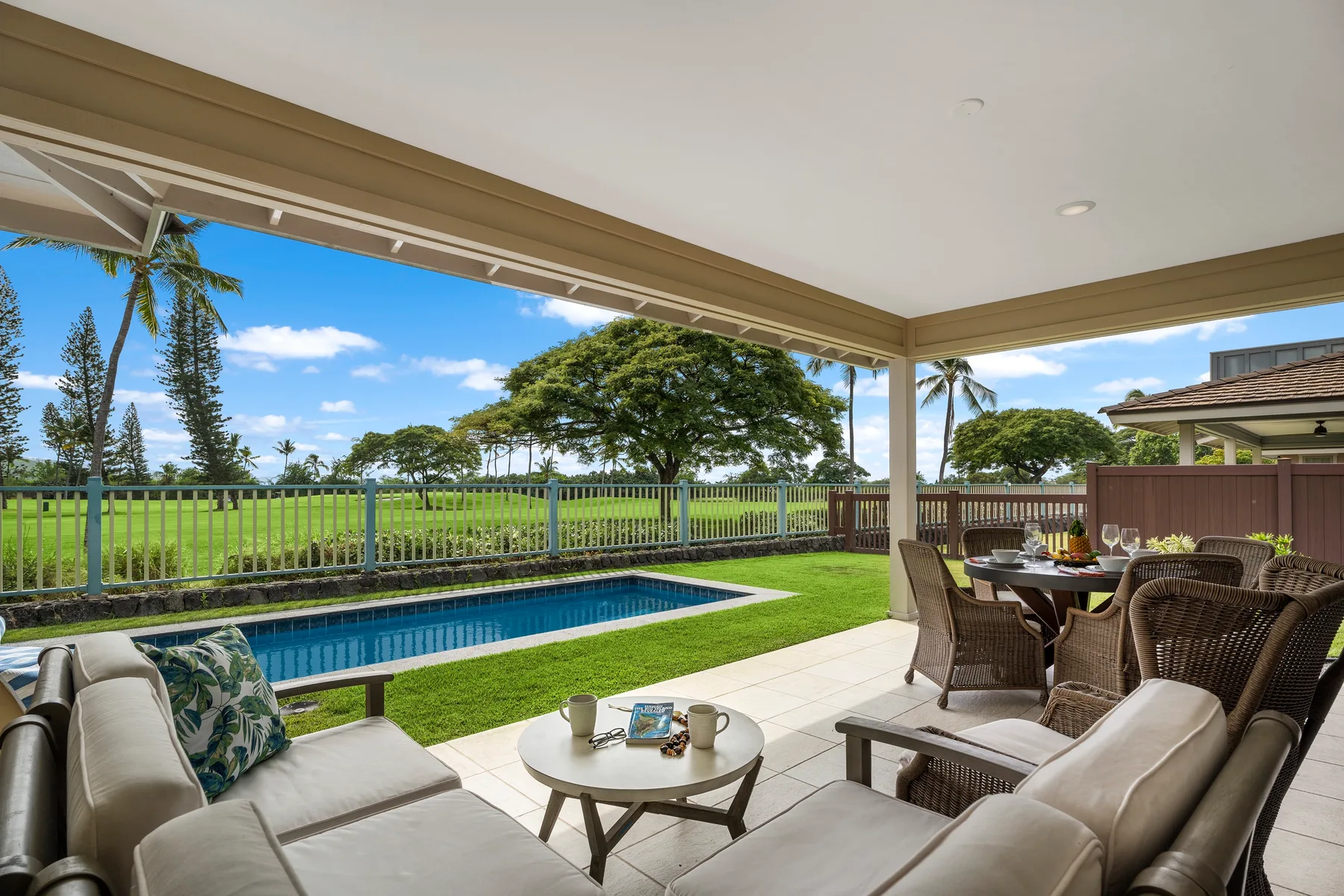 Un patio cubierto con asientos acolchados y una mesa de comedor da a una piscina y un césped.