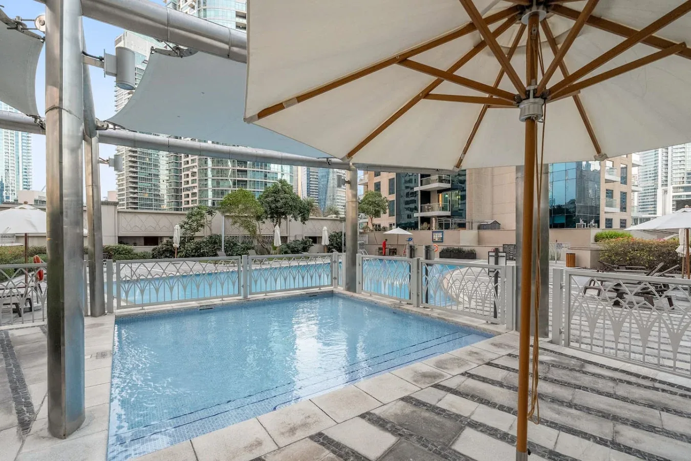 An outdoor swimming pool sits beneath a large shade umbrella beside modern buildings.