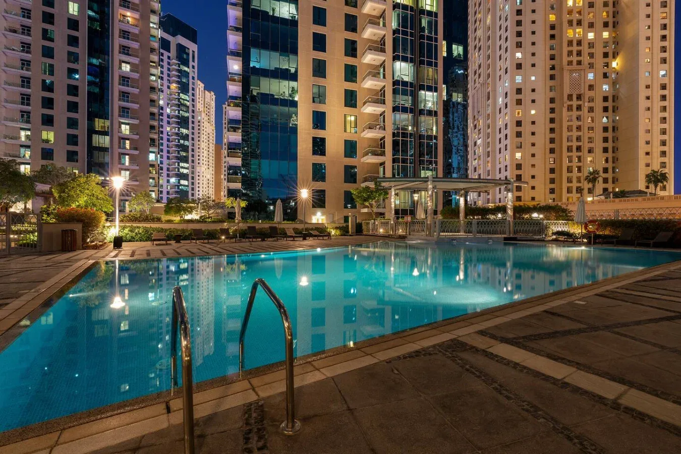A swimming pool glows at night beneath surrounding high-rise buildings.