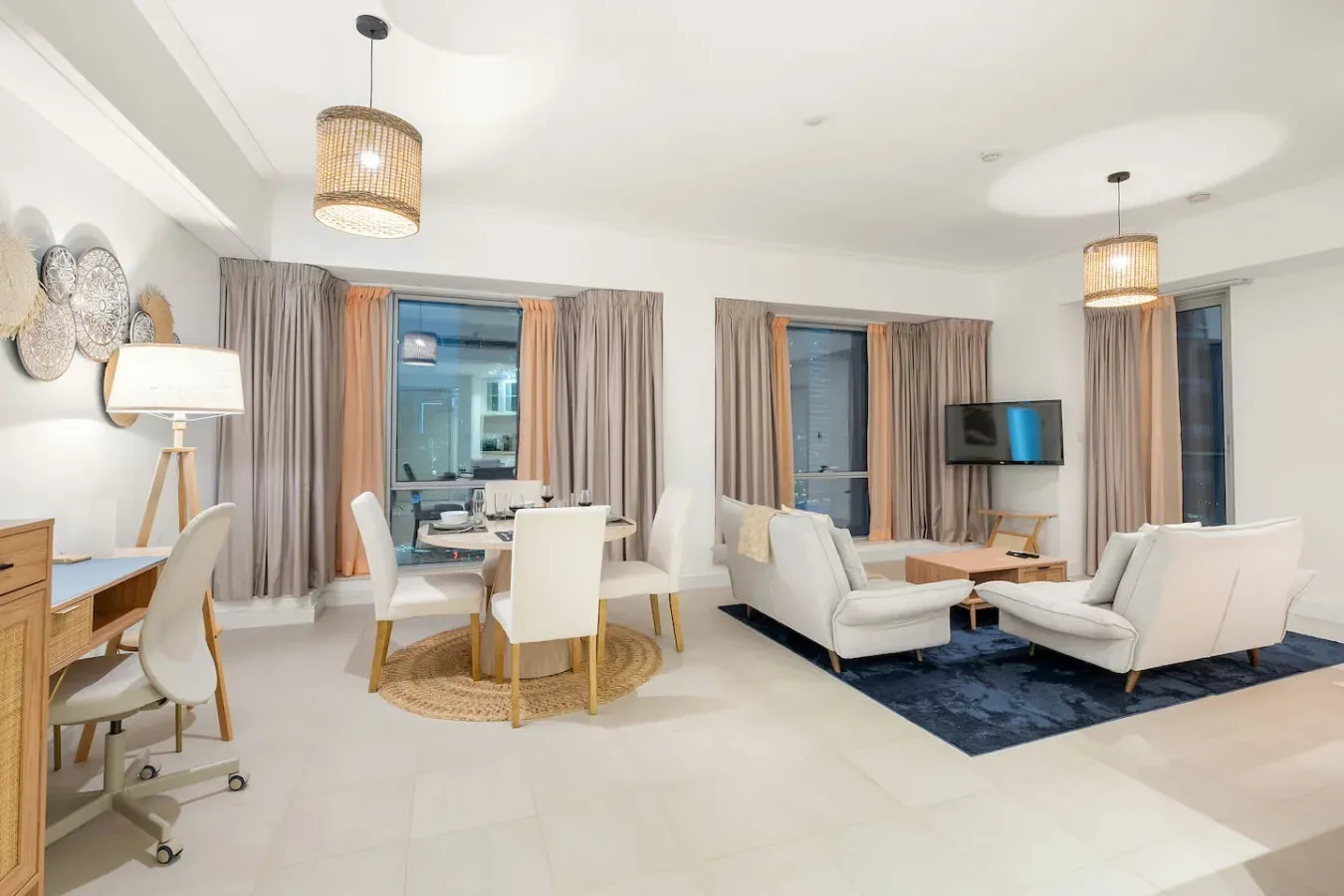A bright living and dining area with seating, a television, and large curtained windows.