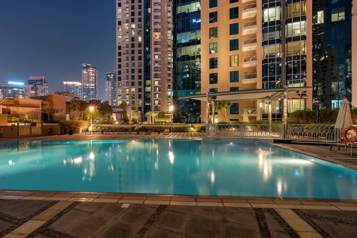 A rooftop swimming pool glows at night among illuminated high-rise buildings.