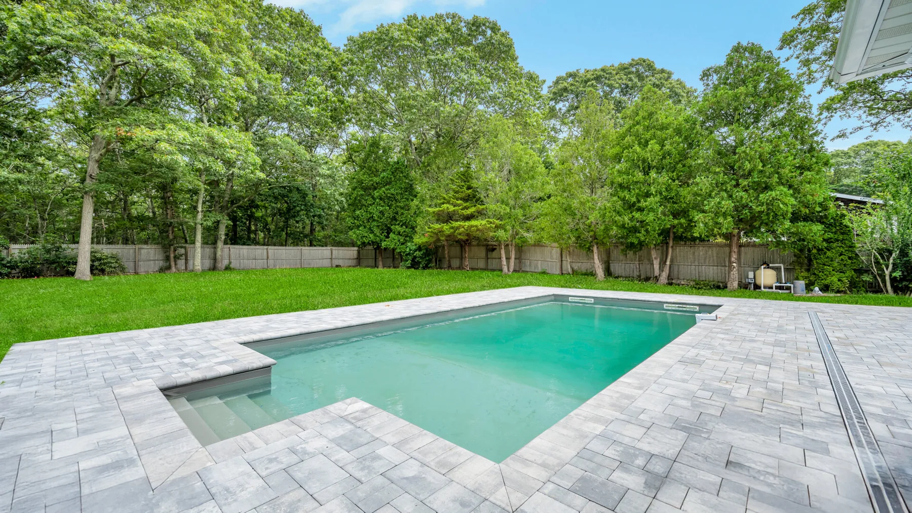 A swimming pool sits in a paved backyard surrounded by trees and a fence.