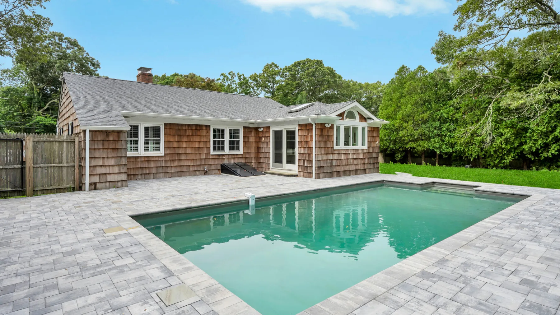 Outdoor swimming pool beside a shingle-sided house, surrounded by a paved patio.