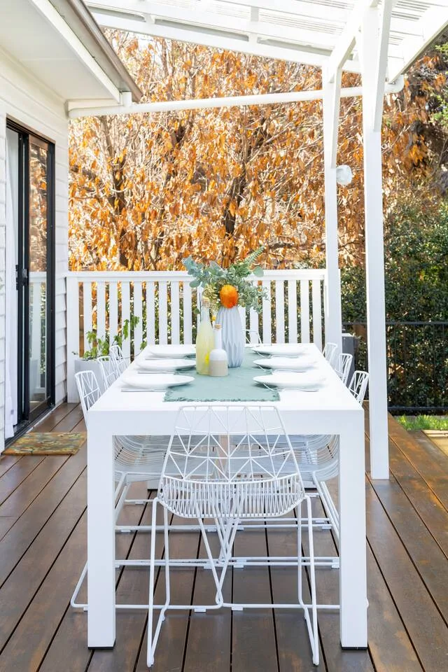 Una zona de comedor exterior cubierta tiene una mesa blanca frente al follaje otoñal.