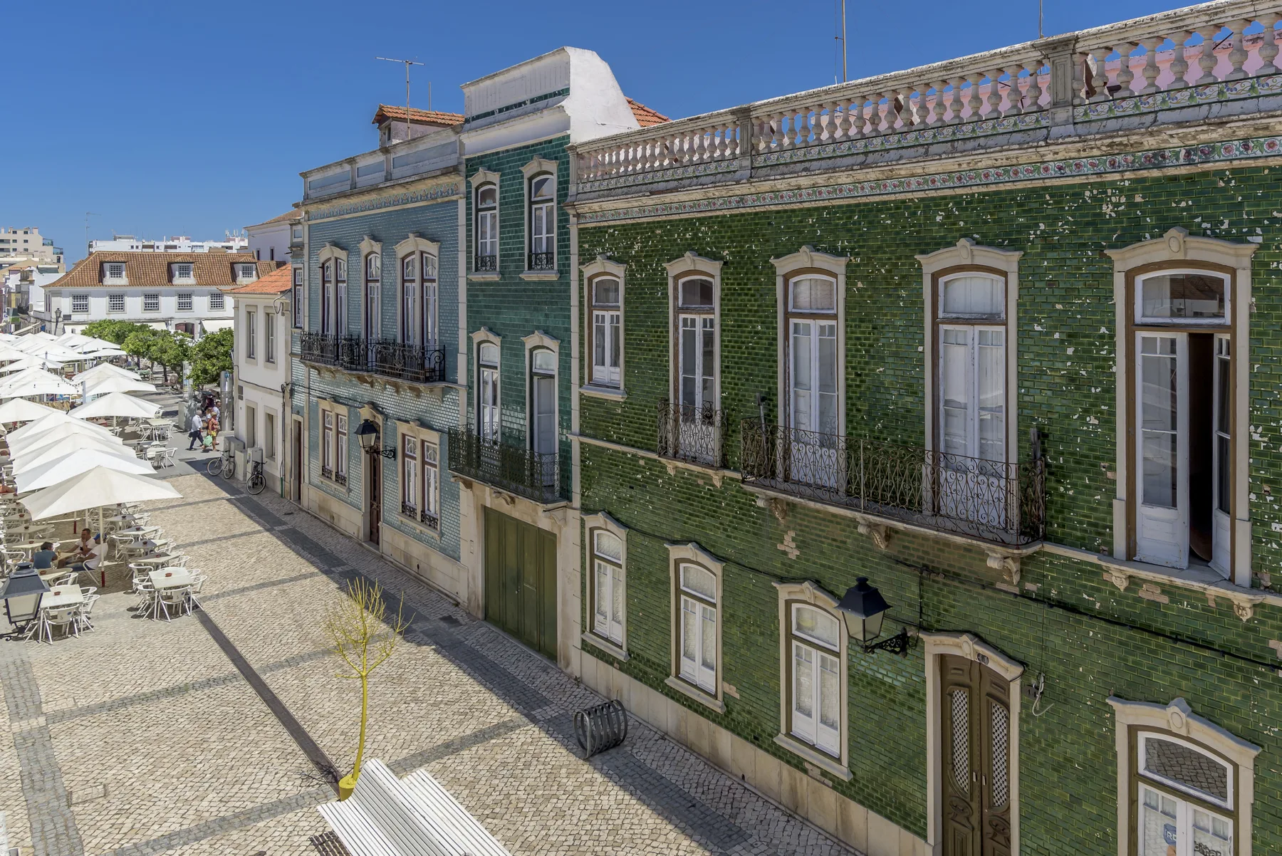 Una calle peatonal empedrada bordea un largo edificio verde con balcones.