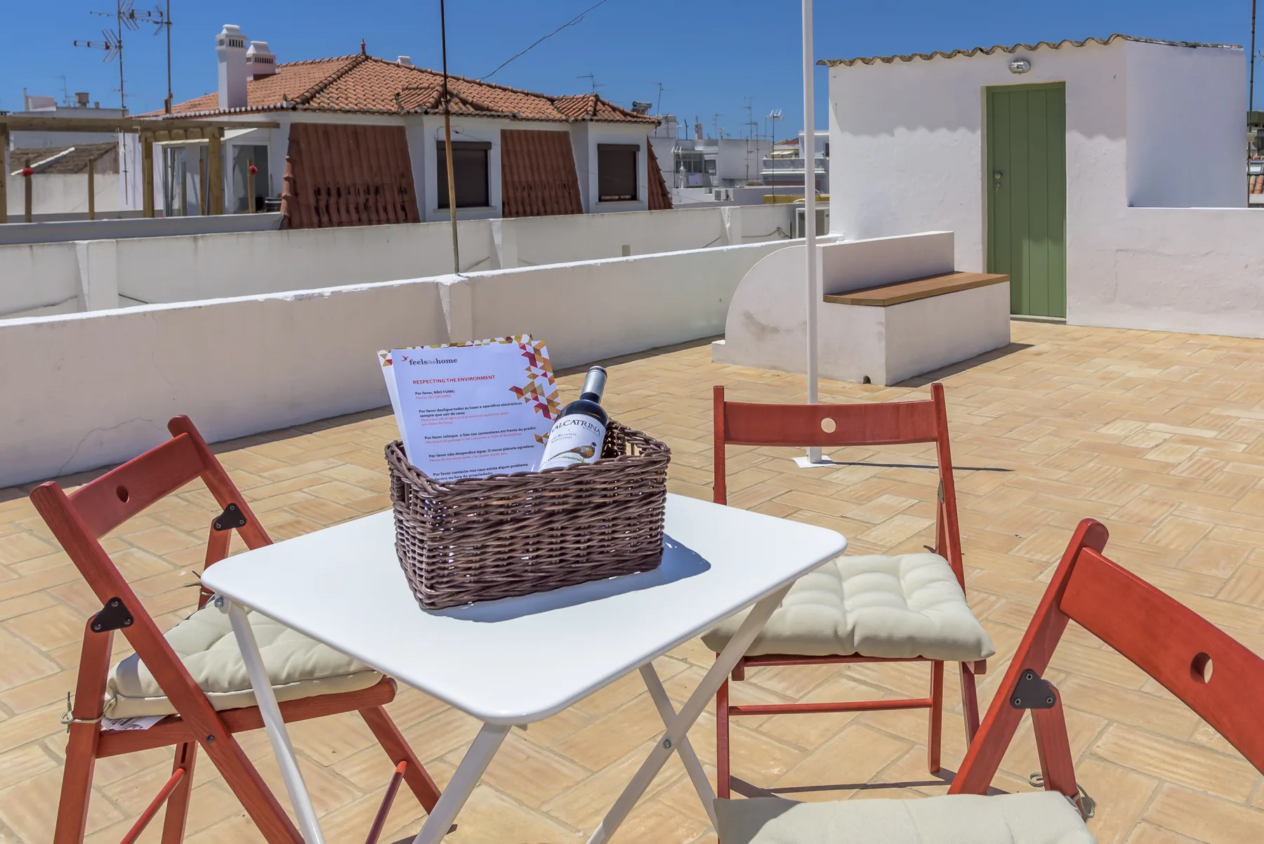Una terraza en la azotea tiene una mesa blanca, asientos de madera y una puerta verde.