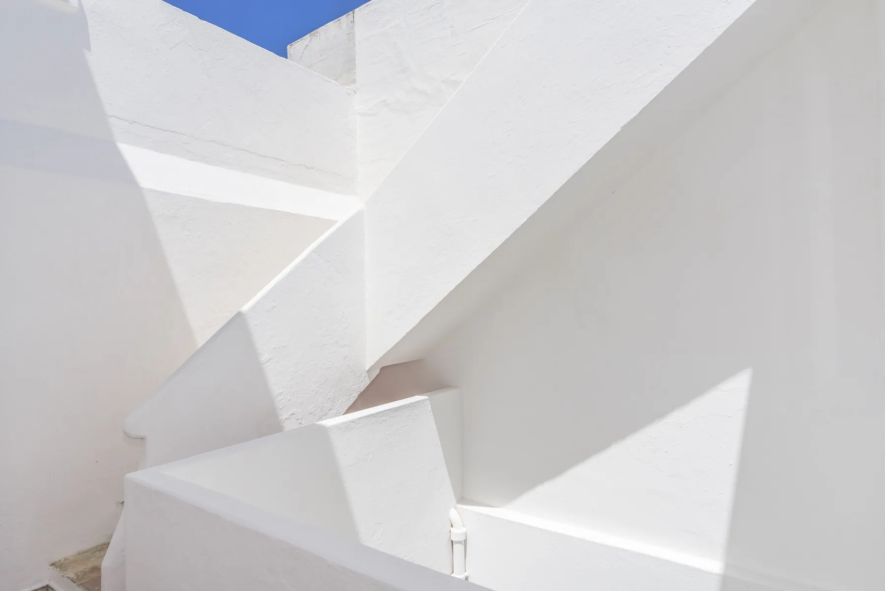 Paredes exteriores blancas y escaleras angulares enmarcan un pequeño tramo de cielo azul.