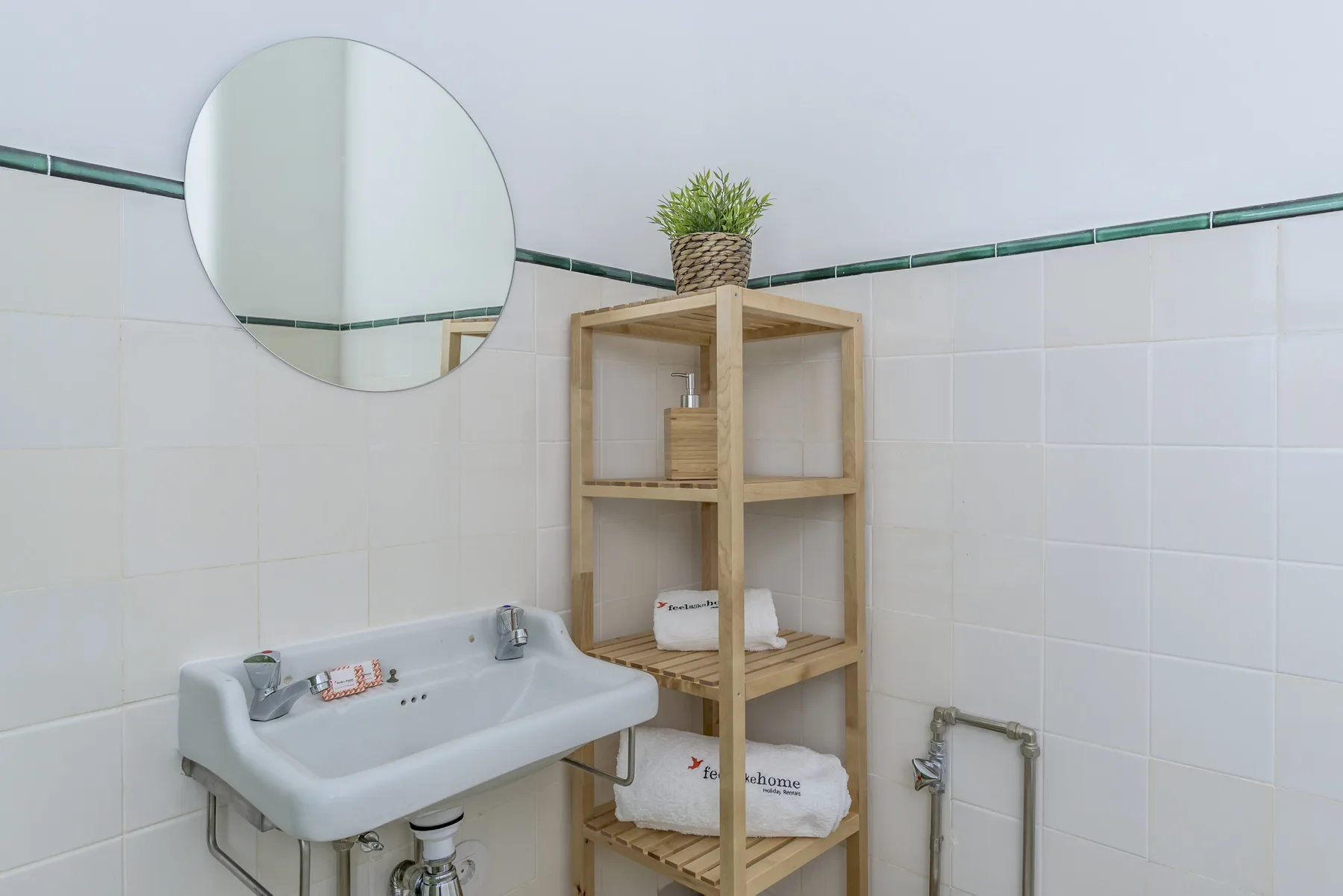 Un baño alicatado con lavabo, espejo redondo y estantería de madera.