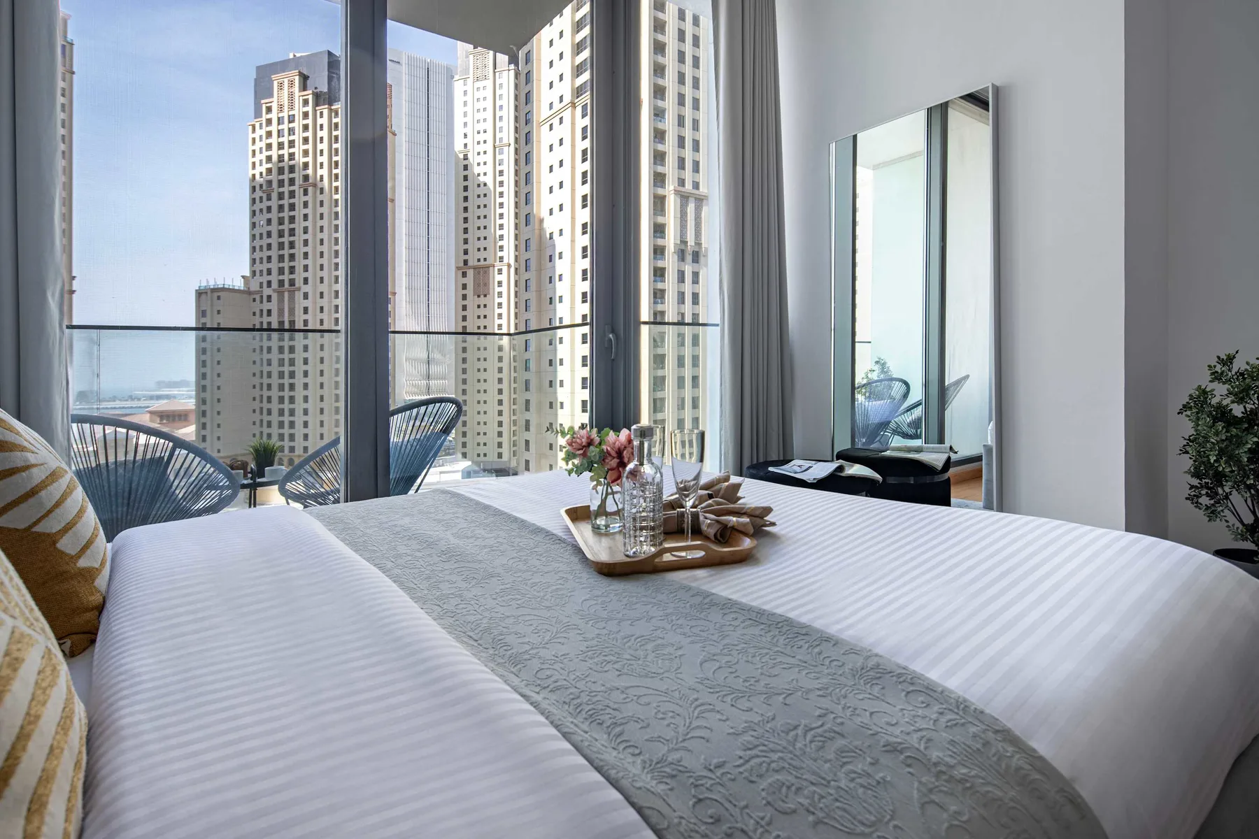 A bedroom with a neatly made bed beside floor-to-ceiling windows overlooking nearby buildings.