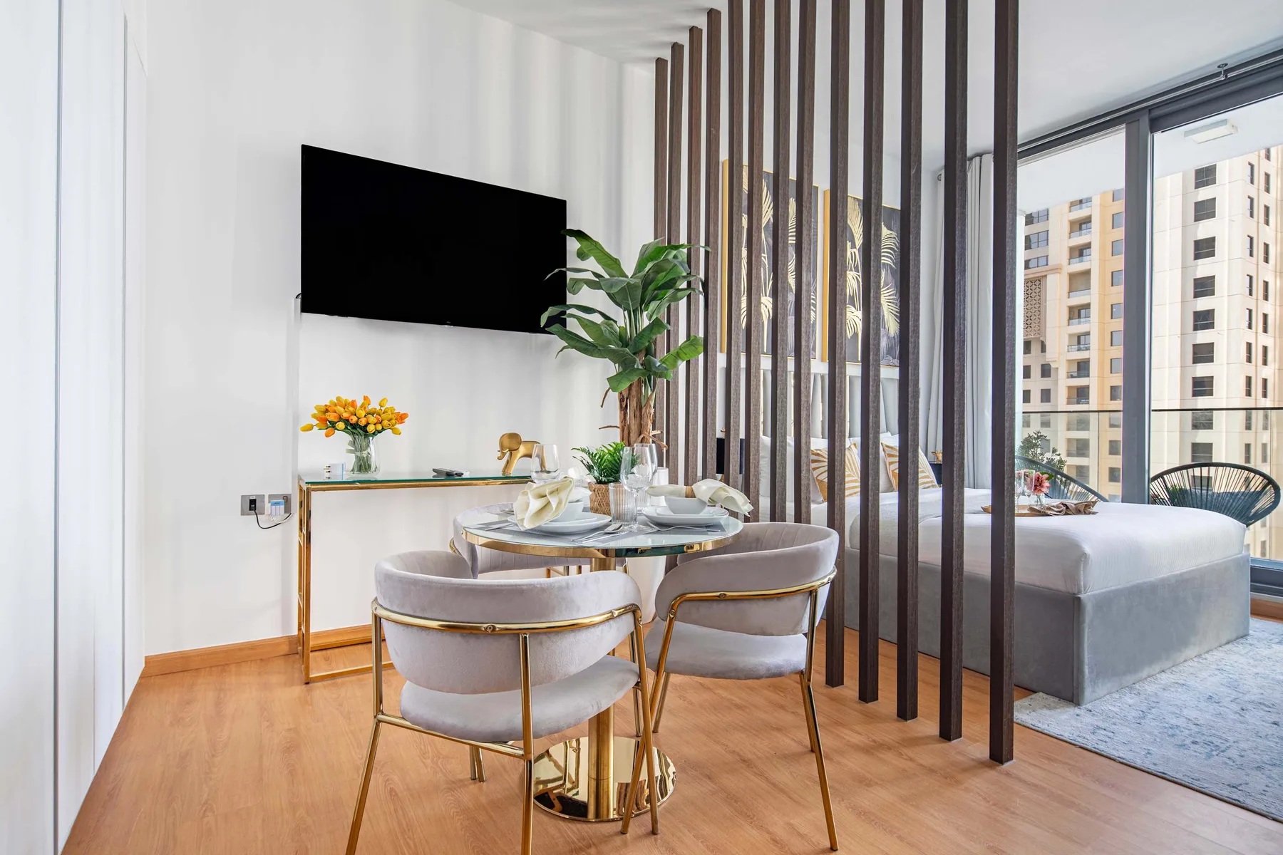 A dining area beside a bed, separated by a slatted divider and overlooking nearby buildings.