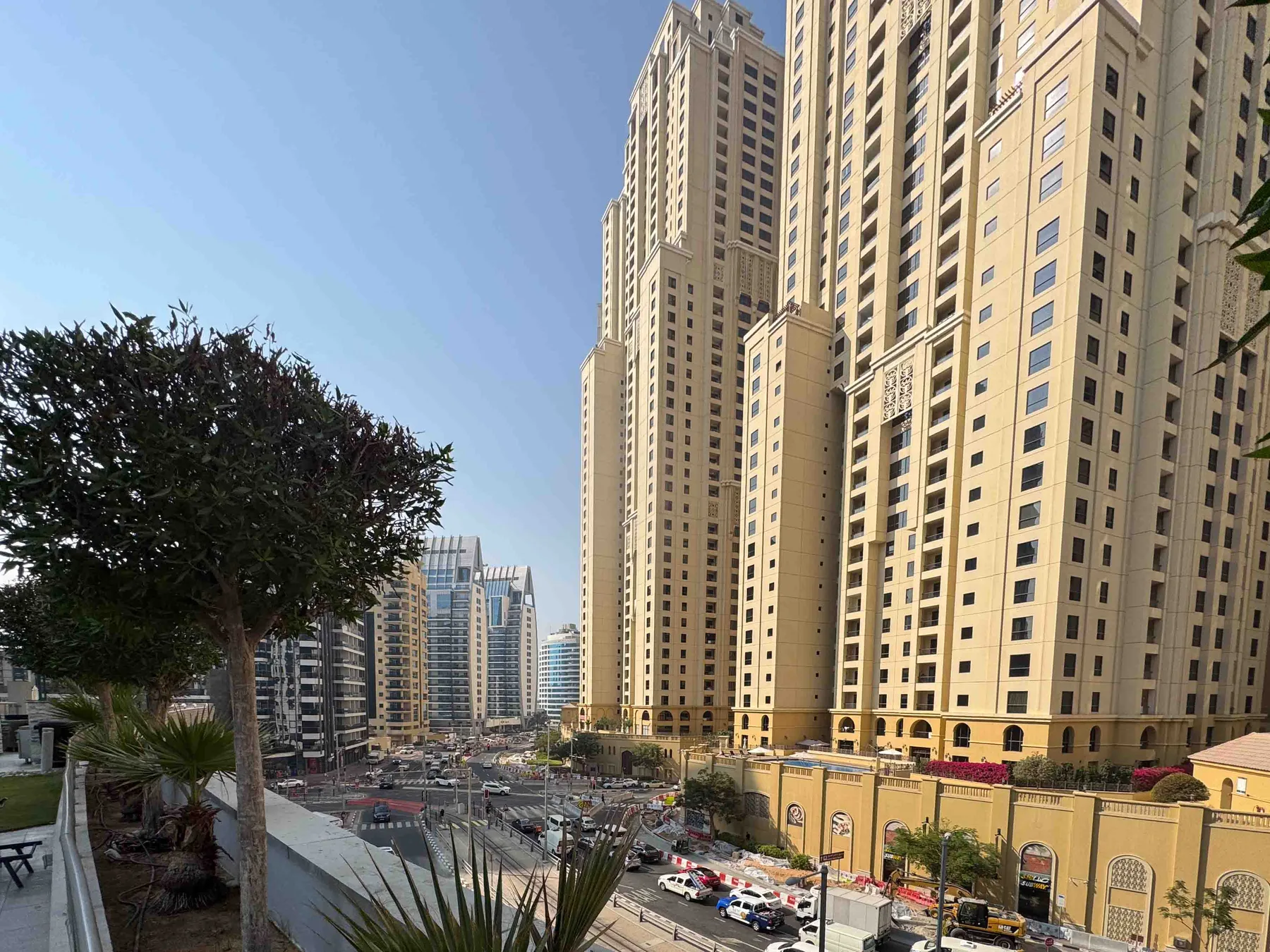 A city street runs beside tall high-rise buildings and a tree-lined sidewalk.
