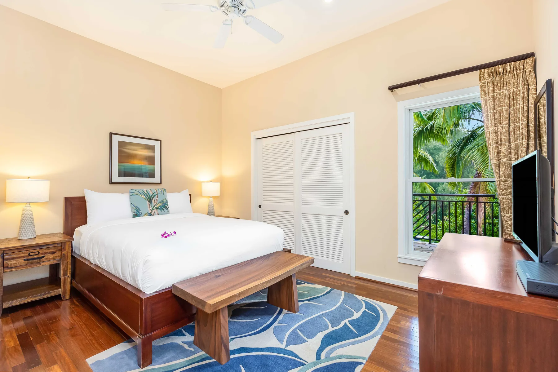 A bedroom with a bed, a blue patterned rug, and a window overlooking palm trees.