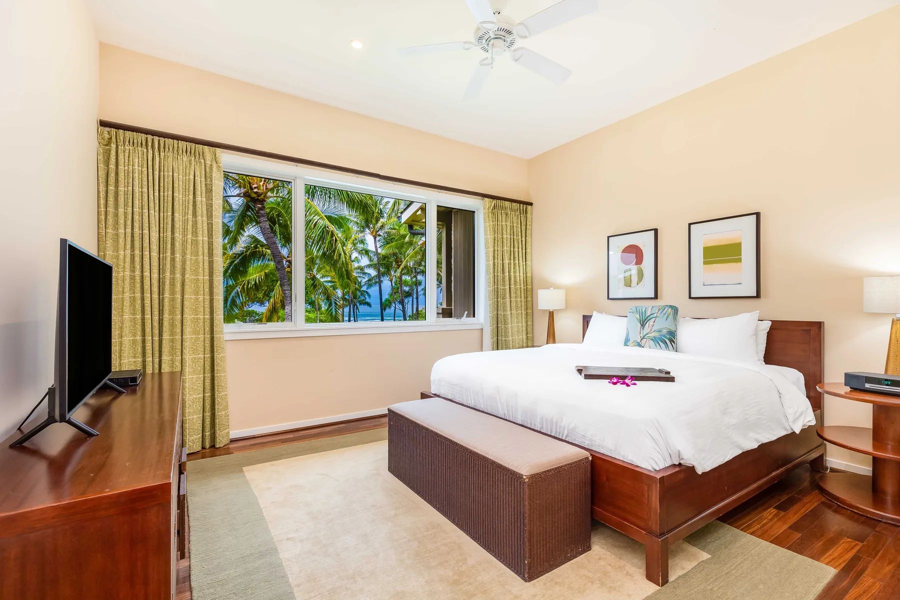 A bedroom with a large bed, bedside tables, and a window overlooking palm trees.