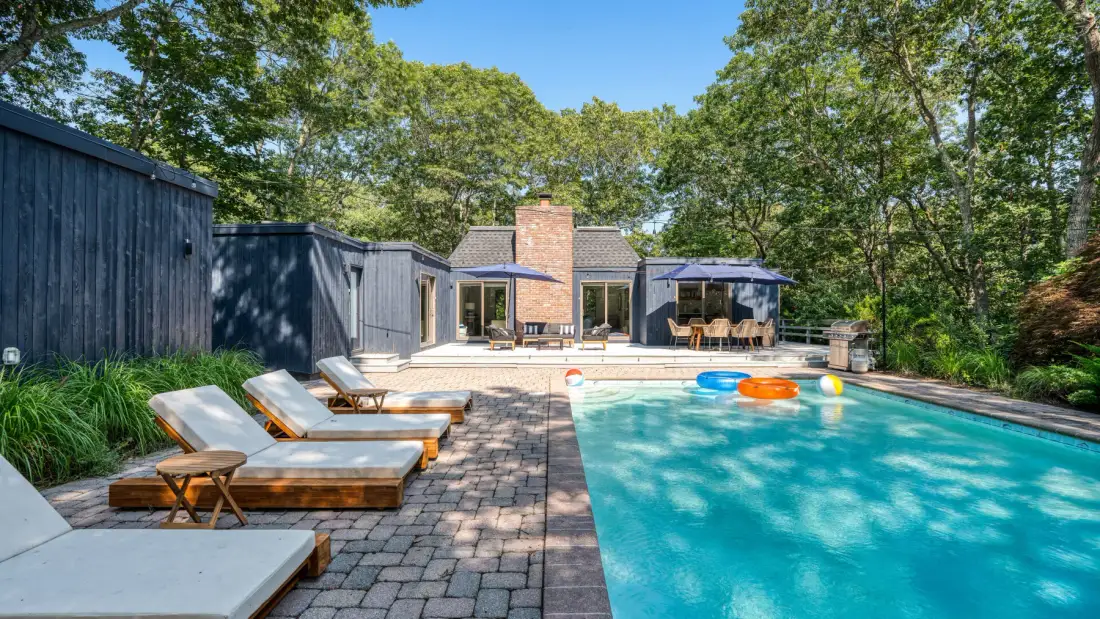 Una piscina al aire libre junto a un patio pavimentado con asientos y árboles.
