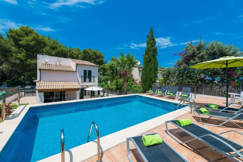 An outdoor swimming pool surrounded by lounge chairs, greenery, and a house.