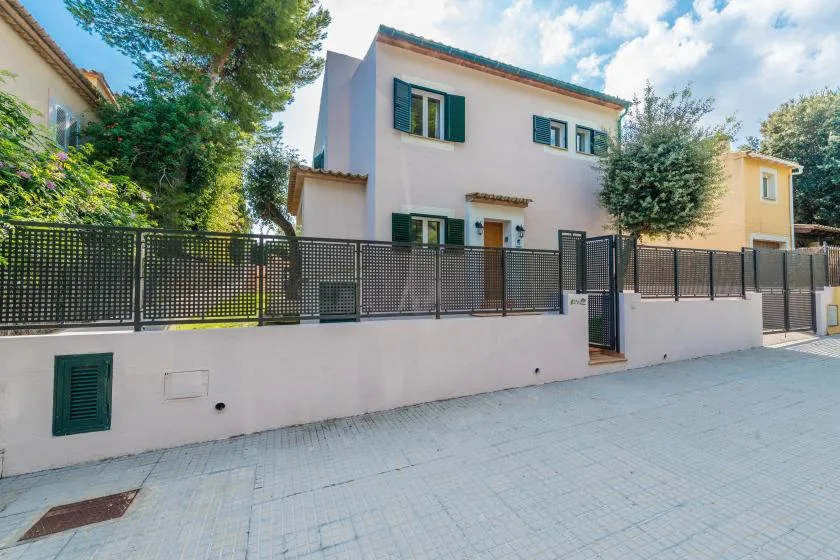 A pale two-story house behind a low wall and black metal fence.
