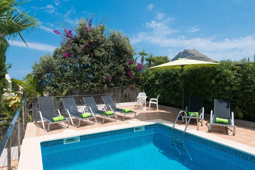 An outdoor swimming pool is surrounded by loungers, greenery, and a sun umbrella.