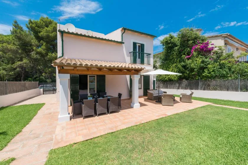 A house opens onto a paved patio with outdoor seating and a green lawn.