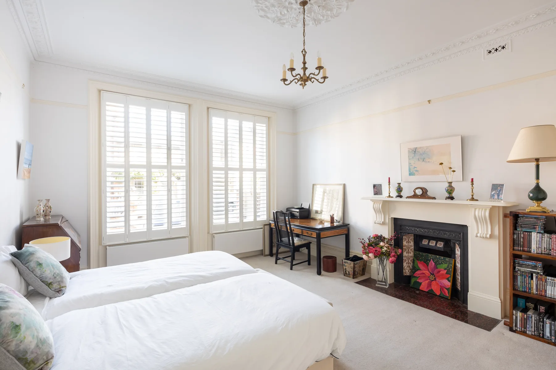A bright bedroom with white bedding, tall shuttered windows, and a fireplace.