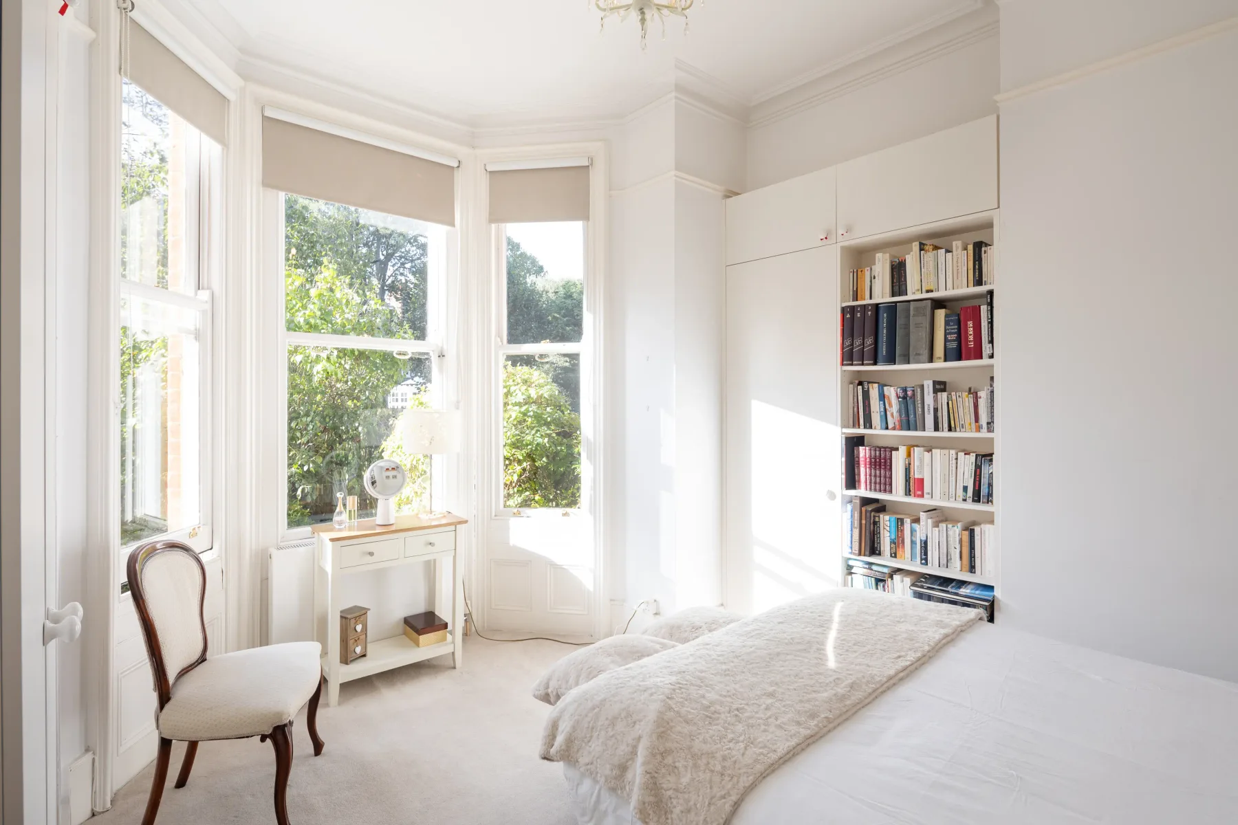 A bright bedroom with tall windows, a bed, and a bookcase.