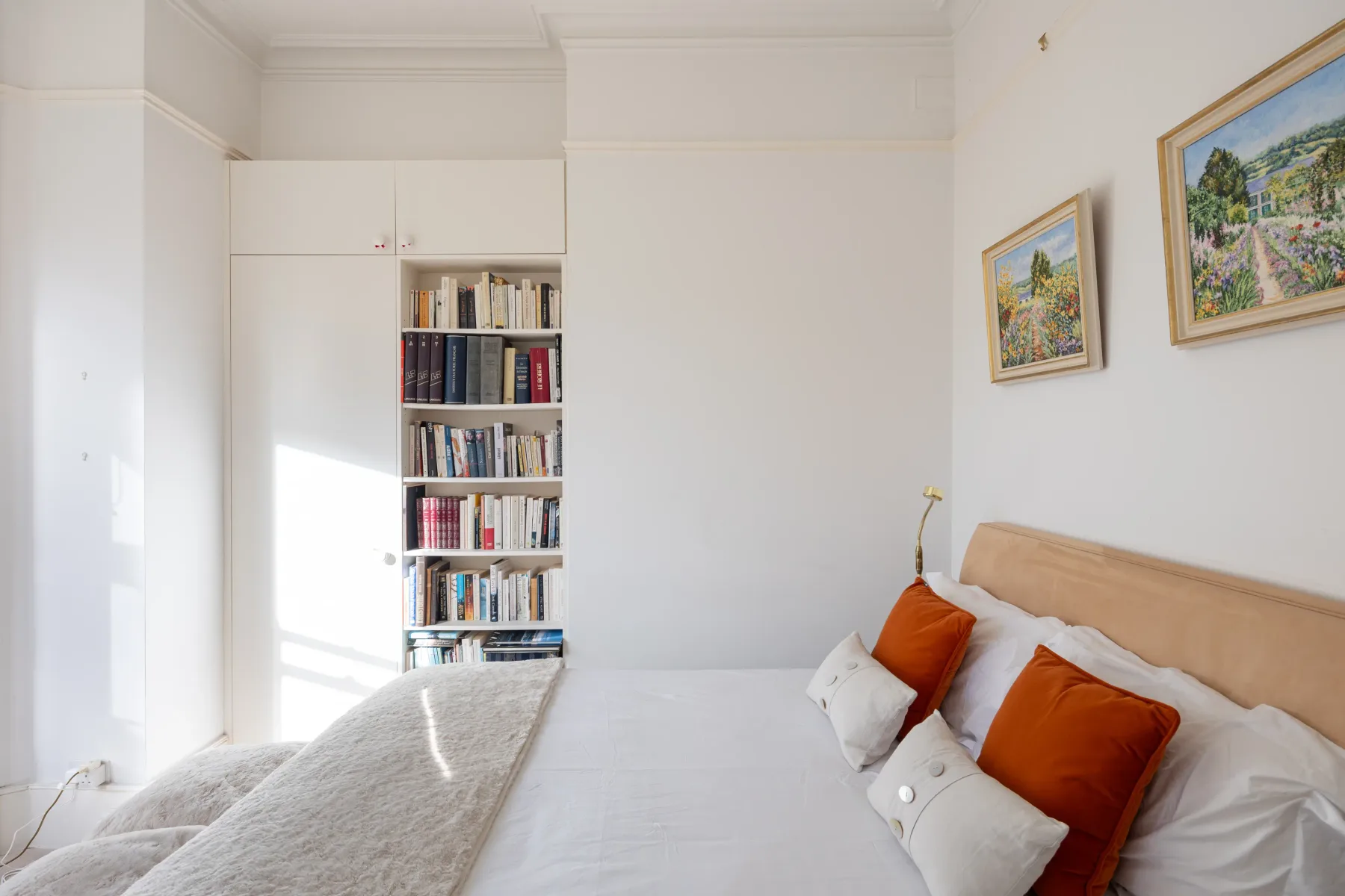 A bright bedroom with a bed, built-in bookshelves, and framed artwork.