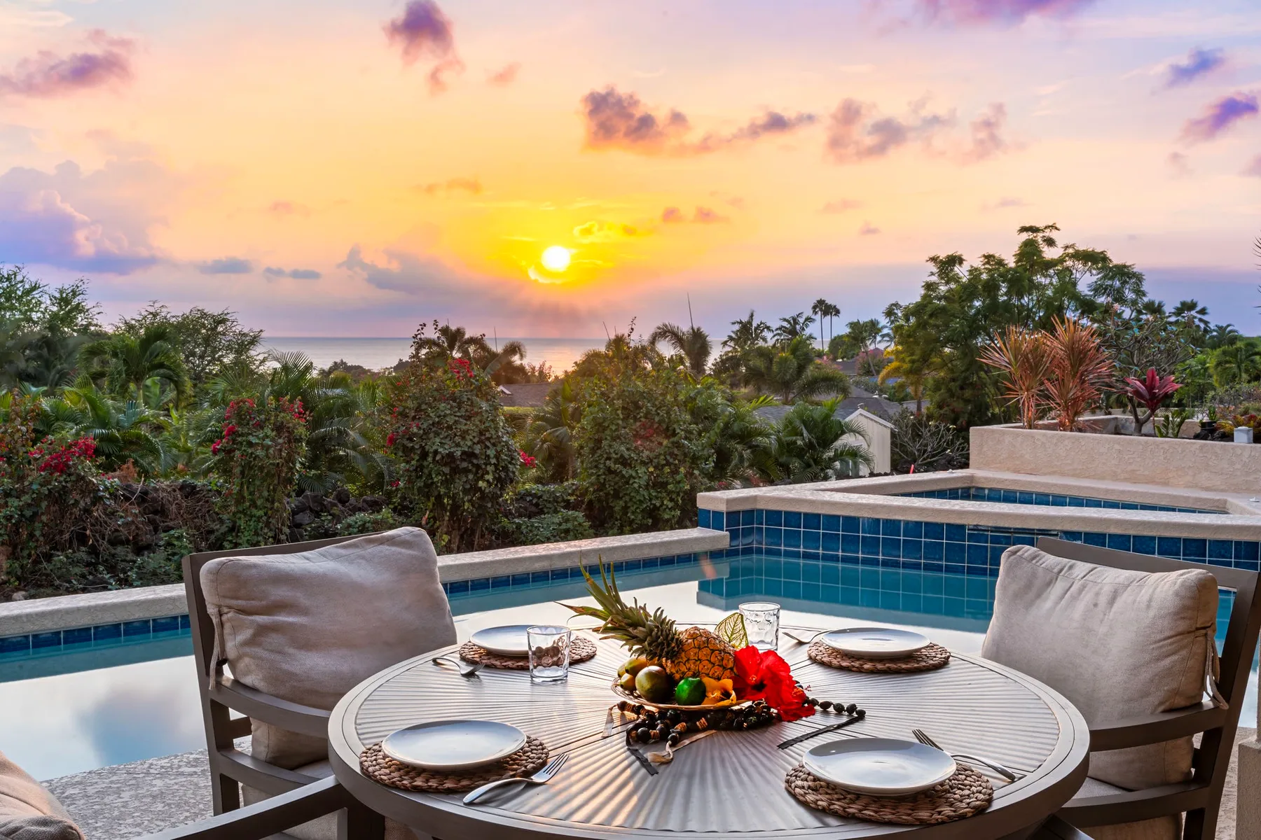 An outdoor dining area beside a swimming pool overlooks lush gardens at sunset.
