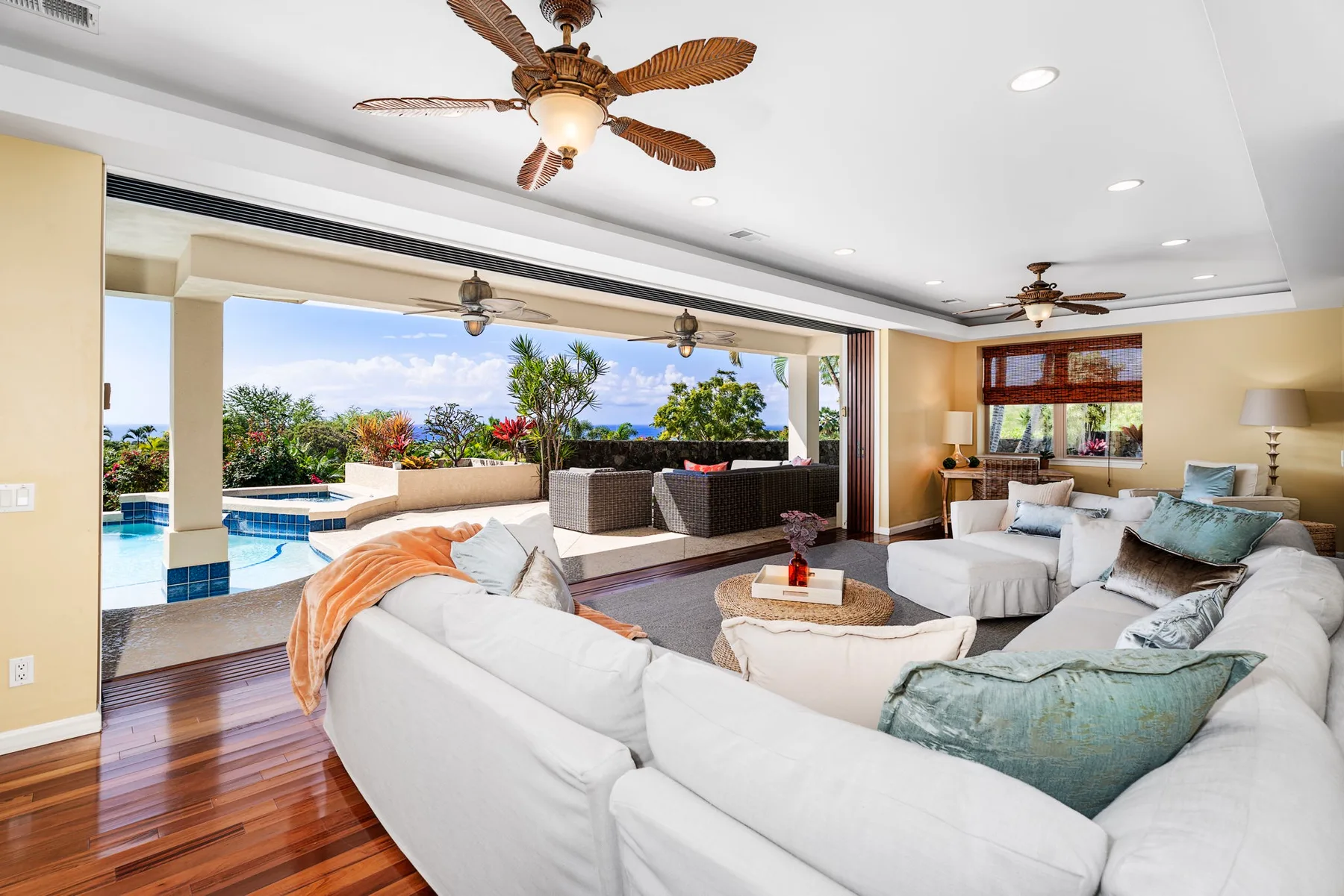 A bright living room opens onto a poolside patio, with white seating and a ceiling fan.
