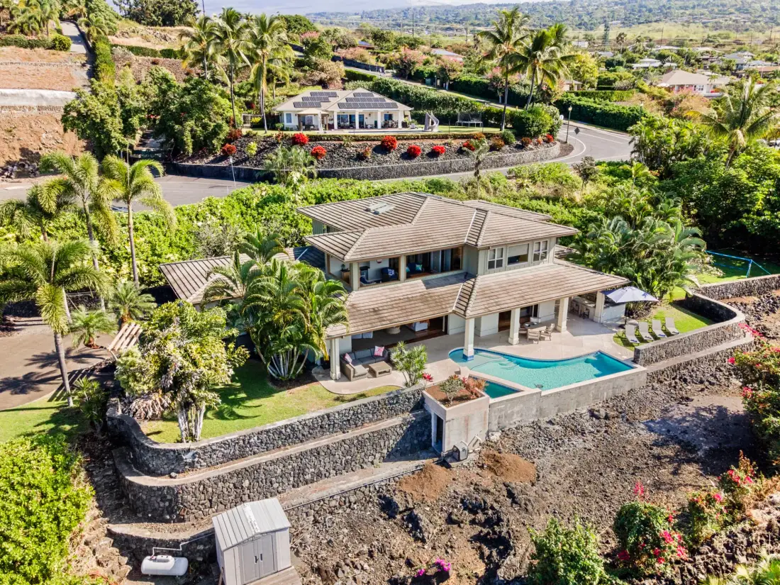 An aerial view shows a hillside house with a swimming pool and landscaped gardens.