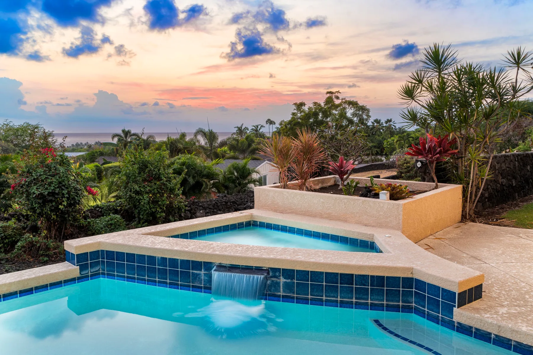 A swimming pool bordered by tropical plants overlooks a colorful sunset sky.