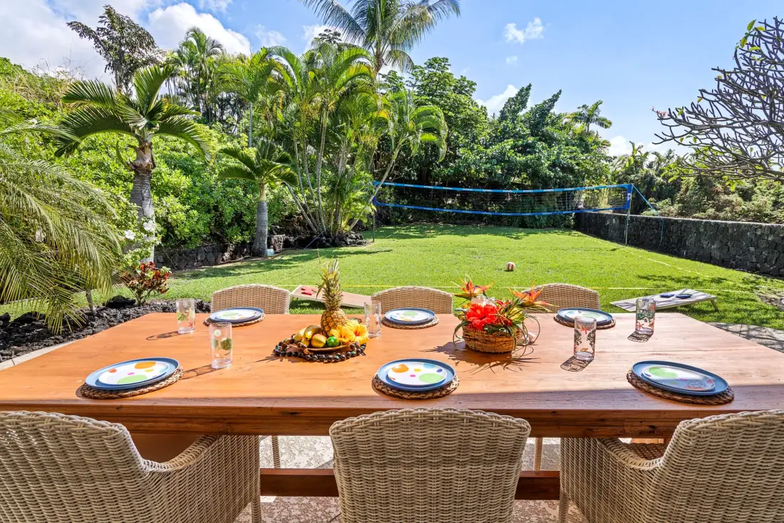 An outdoor dining table is set beside a green lawn and tropical plants.
