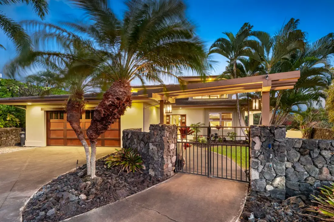 A modern house entrance with a metal gate, stone walls, and palm trees.