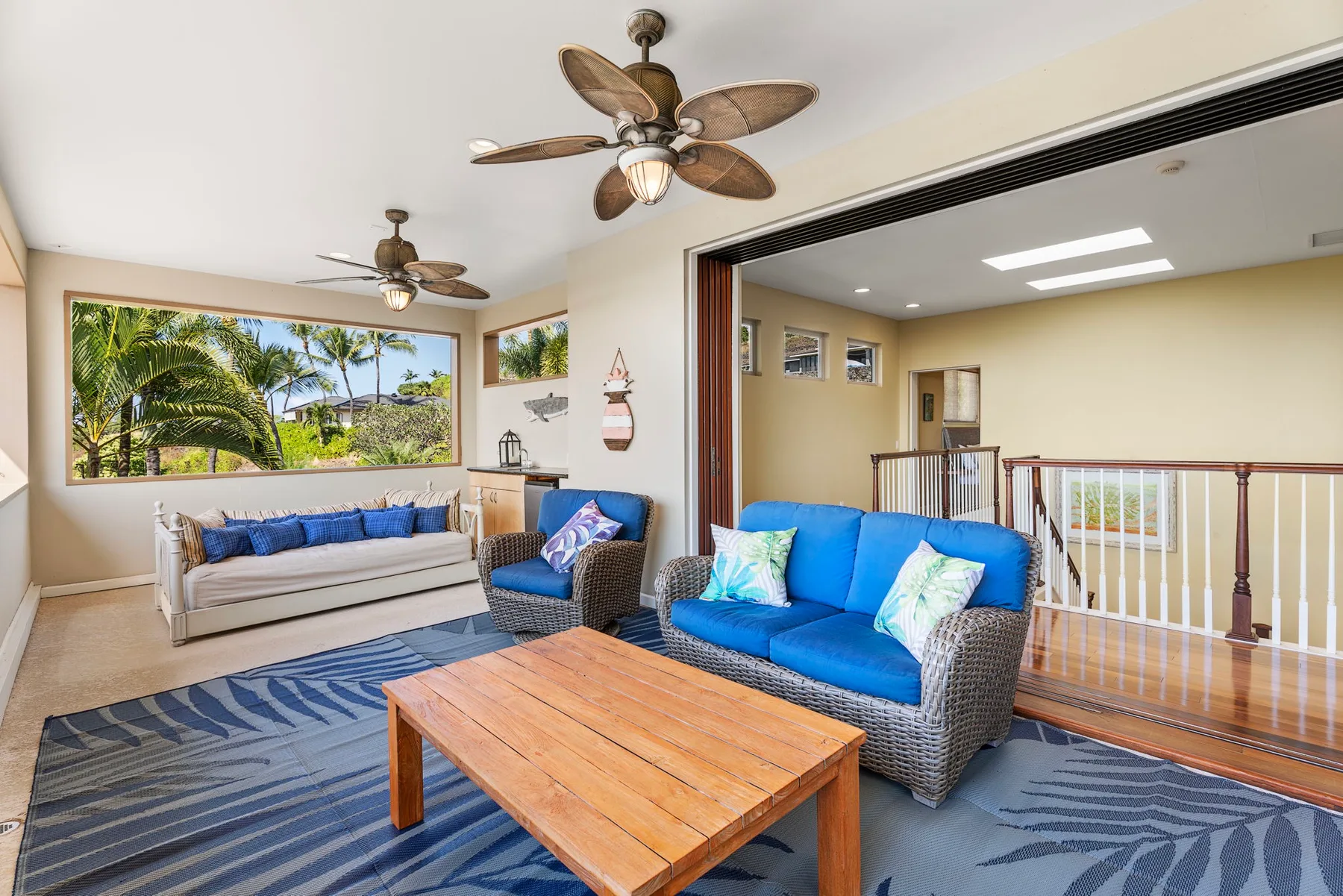 A bright living room with blue seating, a wooden coffee table, and windows overlooking palm trees.