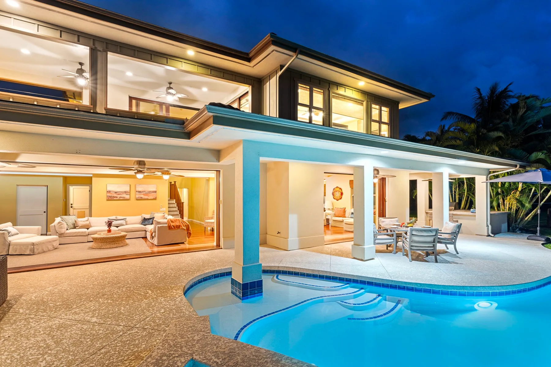 A modern house opens onto a swimming pool and a furnished outdoor patio at dusk.