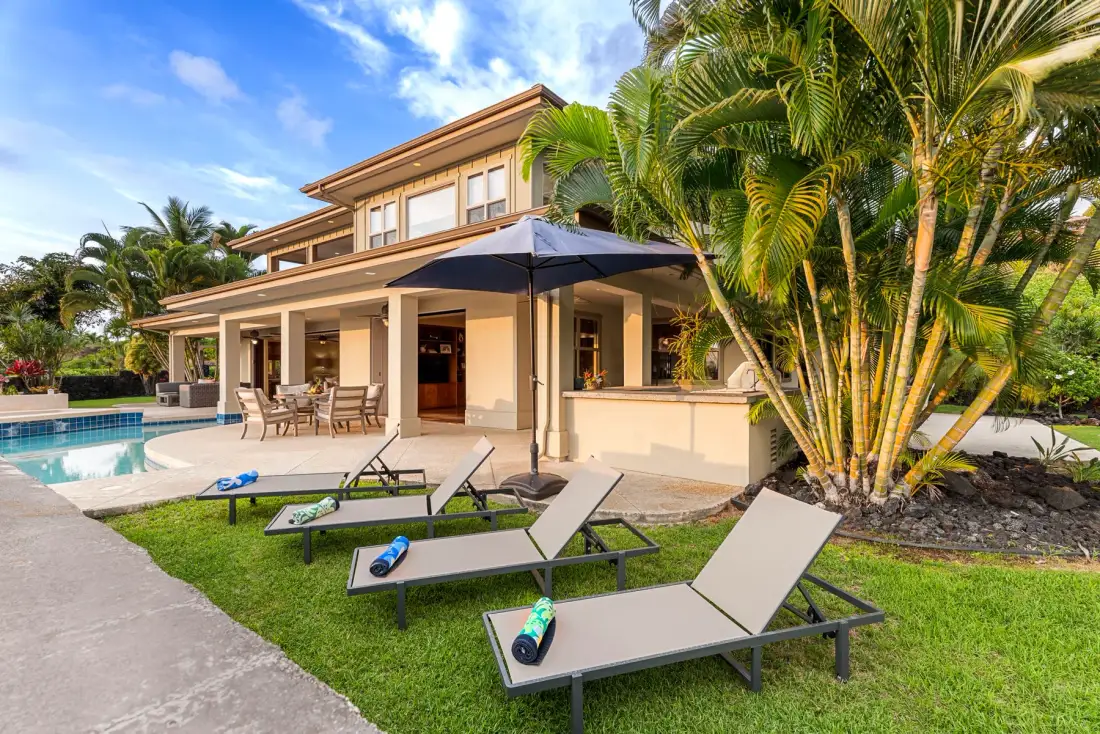 A tropical house beside a swimming pool, with lounge chairs on the lawn.