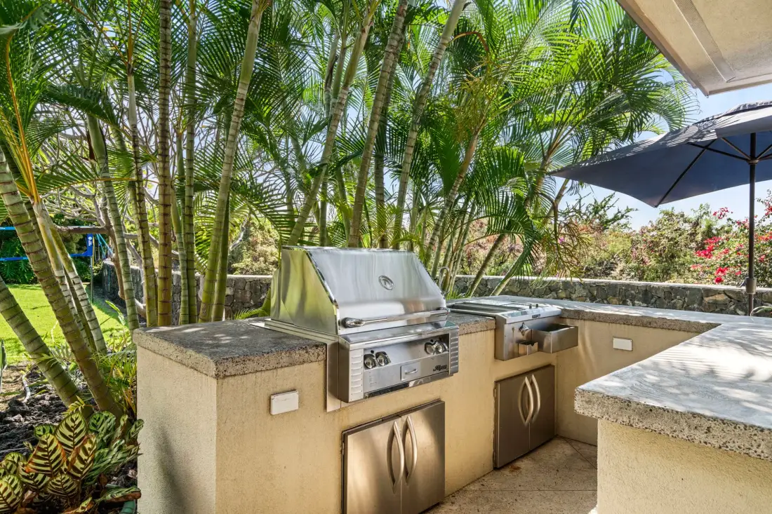 An outdoor kitchen with a built-in grill and countertop, surrounded by palm trees.