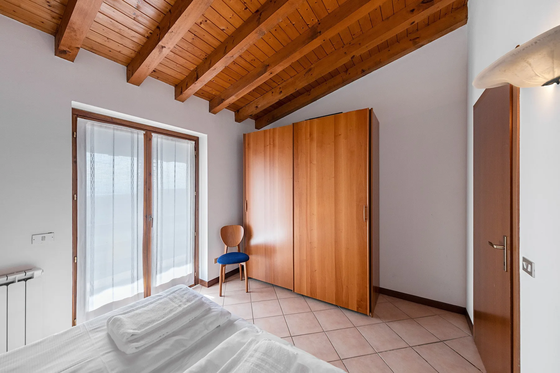 A bedroom with a wooden wardrobe, a chair, and a sloped timber ceiling.