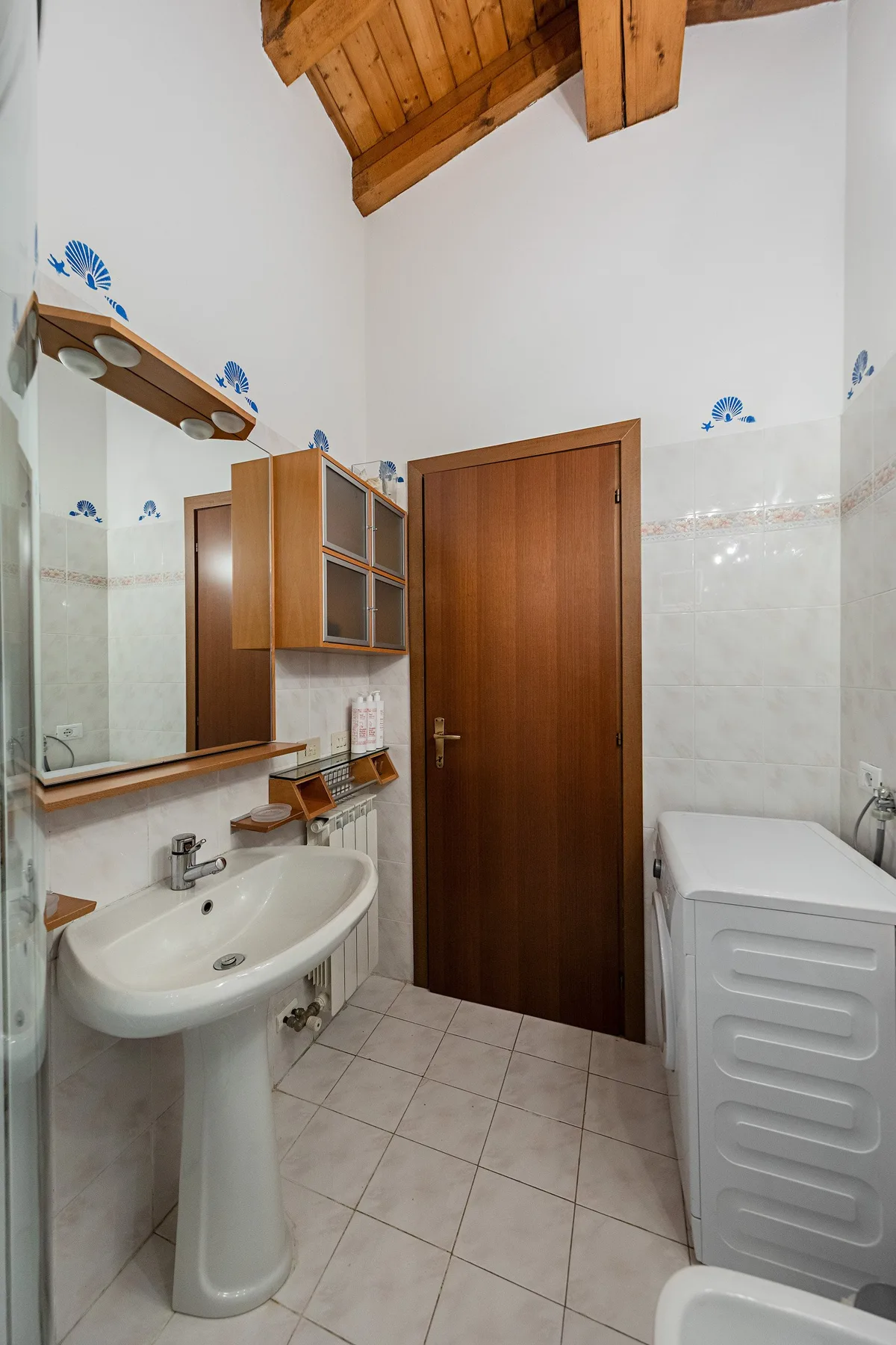 A tiled bathroom with a pedestal sink, wooden door, and washing machine.
