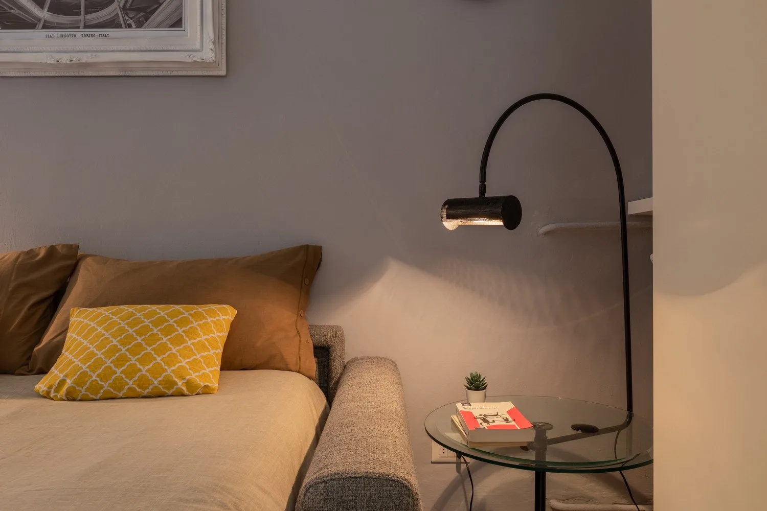 Una zona de dormitorio con una cama, cojines y una lámpara de lectura junto a una mesa de vidrio.