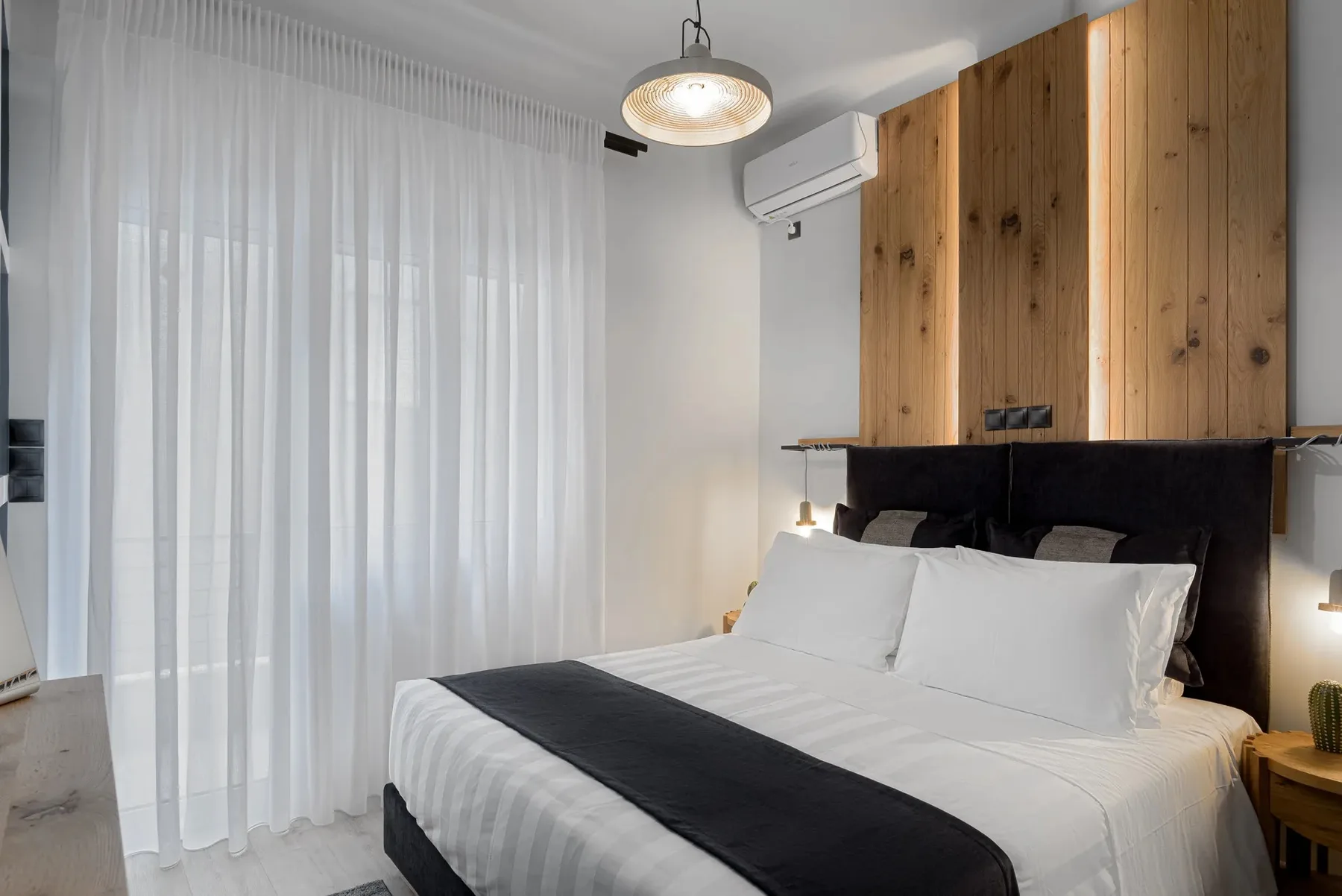 A bedroom with a neatly made bed, sheer curtains, and a wood-paneled wall.