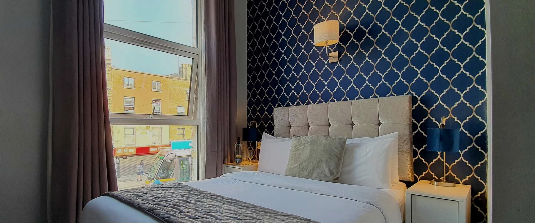 Habitación de hotel con una cama, papel tapiz azul, una ventana grande y lámparas de noche.
