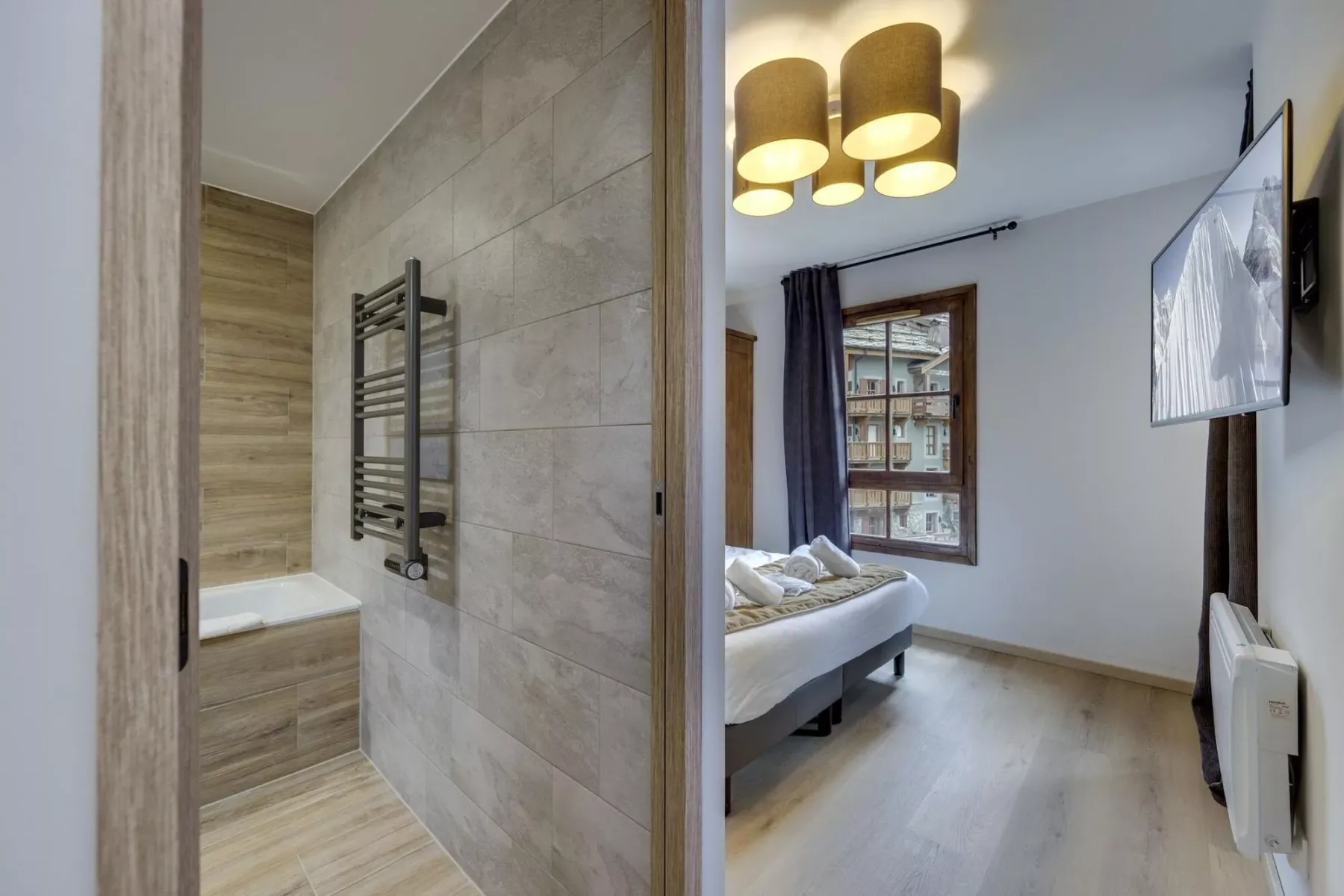 A bedroom with a bed, window, and wall-mounted television beside an open bathroom.