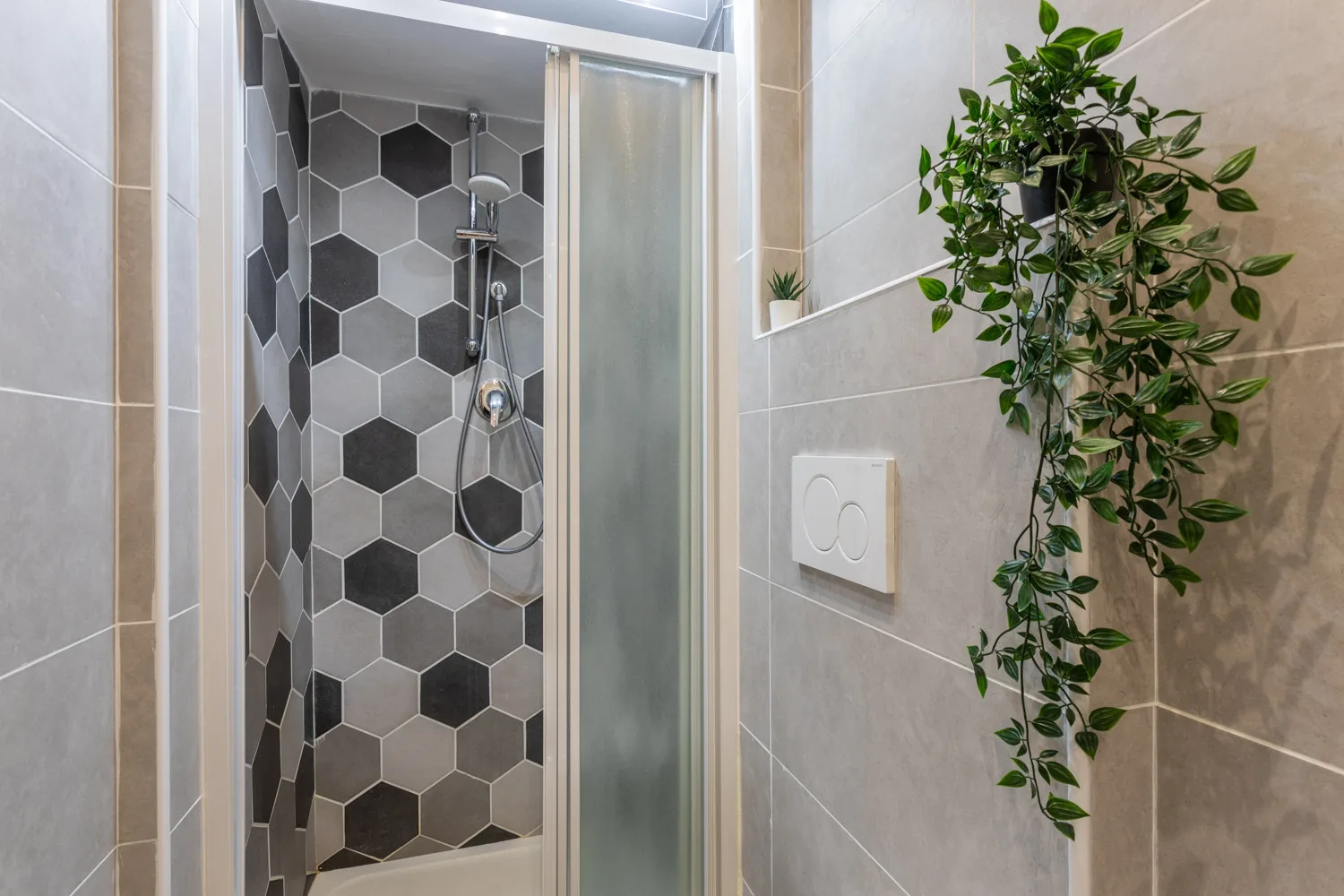 A tiled bathroom shower with a handheld showerhead and a leafy plant.