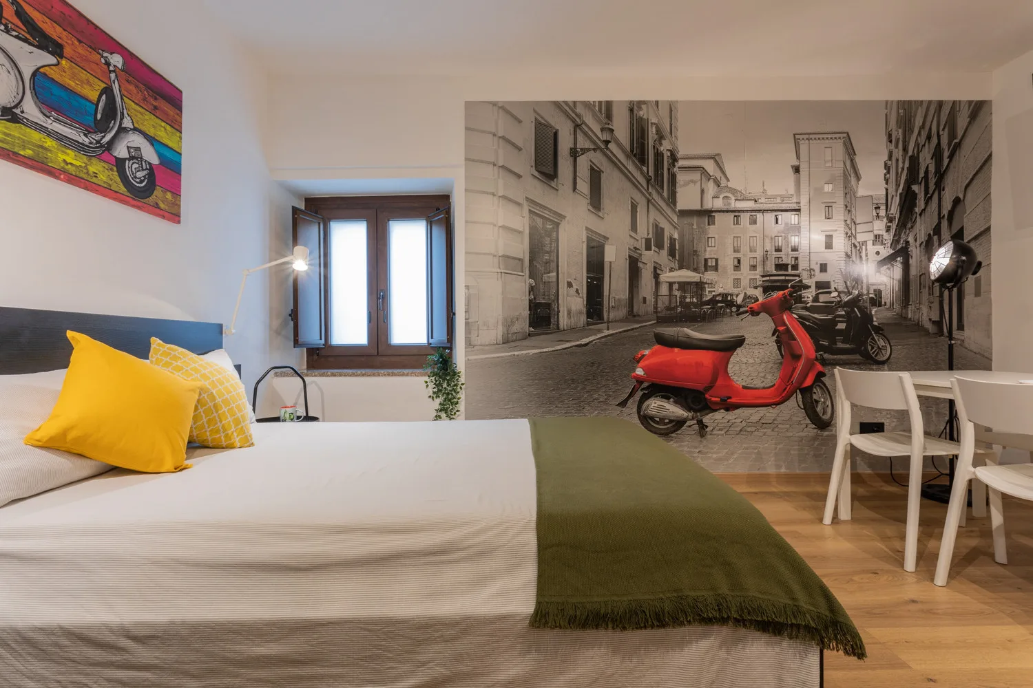 A bedroom with a bed, a window, and a dining table beside a city-street mural.