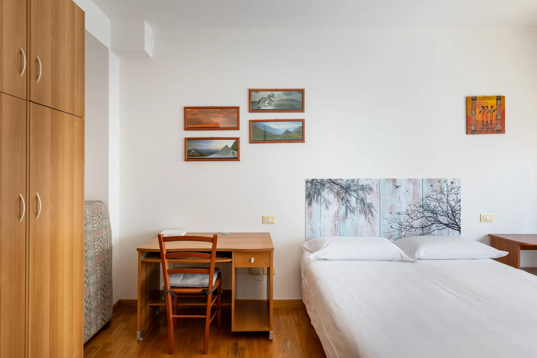 A bedroom with a bed, a desk, and framed artwork on the walls.
