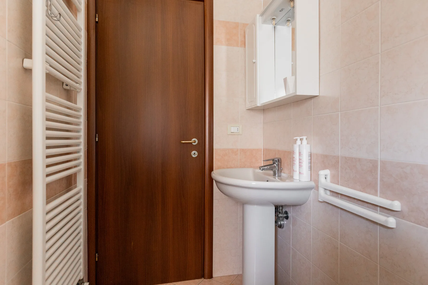 A tiled bathroom with a pedestal sink, wall mirror, and grab bars.