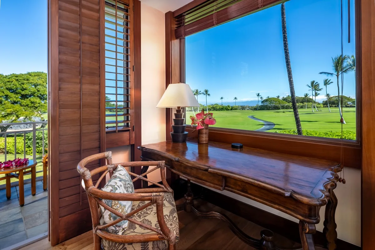 A wooden desk and chair sit beside a wide window overlooking a green landscape.