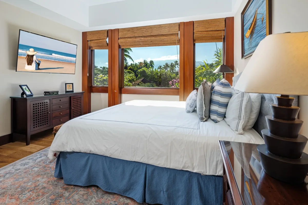 A bedroom with a neatly made bed and windows overlooking tropical greenery.