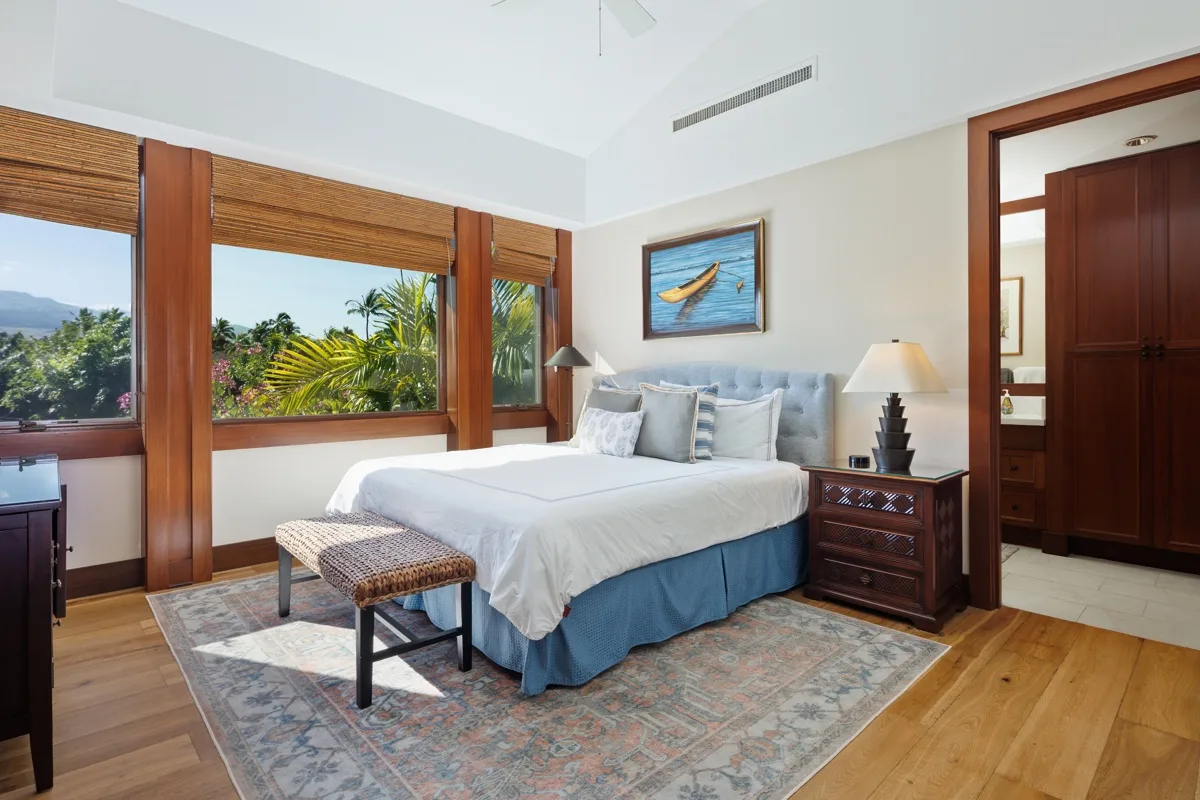A bedroom with a neatly made bed, patterned rug, and large windows overlooking greenery.