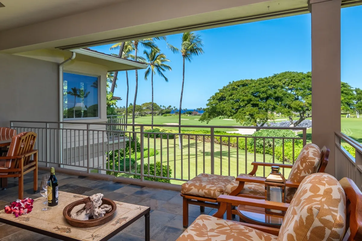 A covered patio with cushioned seating overlooks a green lawn and palm trees.